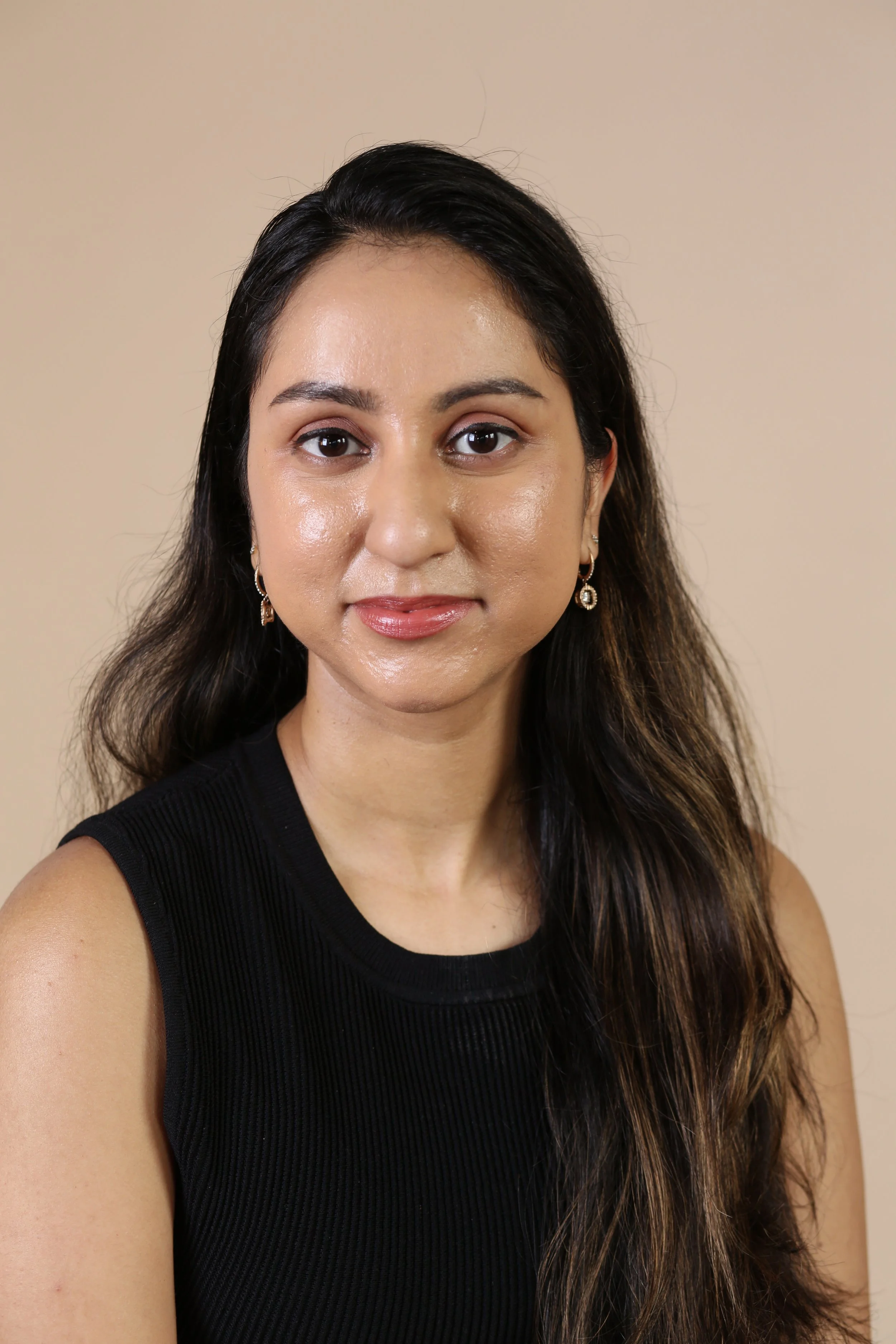 A south asian anxiety people pleasing therapist in New York City with long dark hair, wearing a sleeveless black top and silver earrings, facing forward against a plain beige background.
