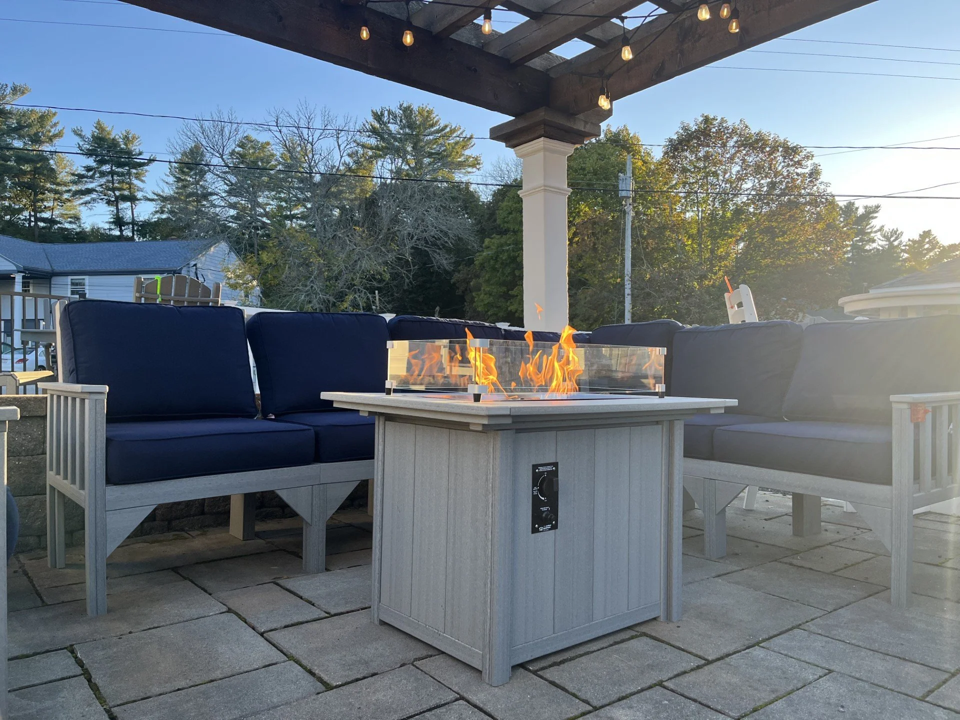 Outdoor patio with cushioned seating around a fire pit, string lights overhead, and trees in the background during daylight.
