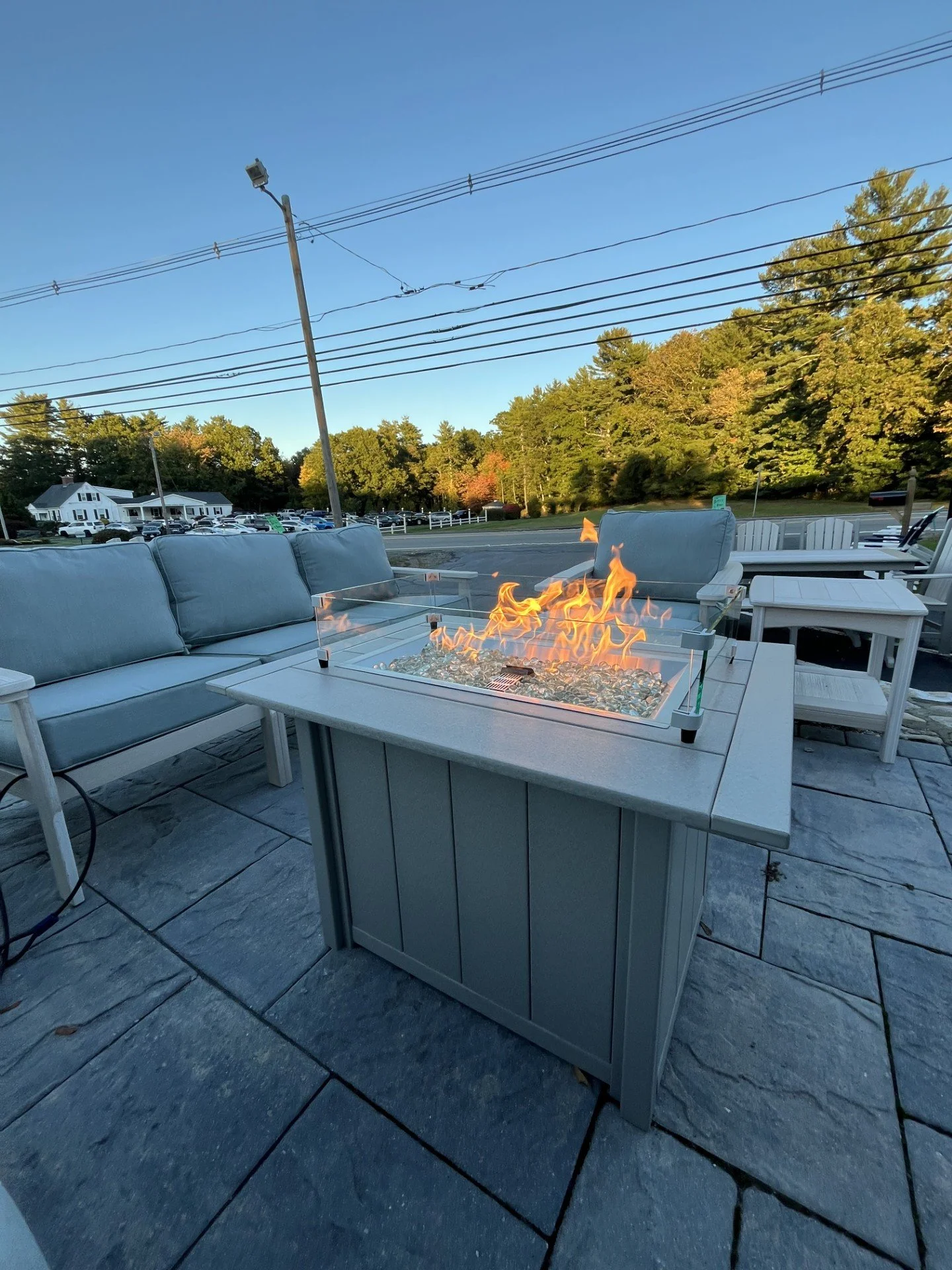 Outdoor patio seating with a fire pit table, cushioned chairs, and tables, with trees and a road in the background during evening time.