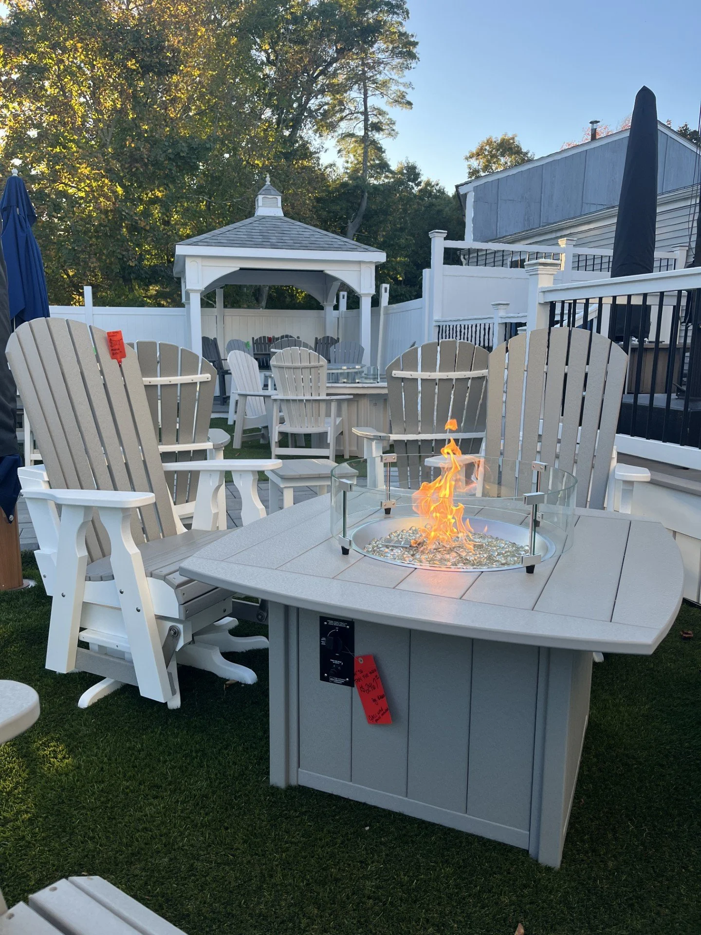 Outdoor backyard patio with a fire pit table surrounded by Adirondack chairs, a gazebo in the background, and other outdoor furniture, fences, and trees