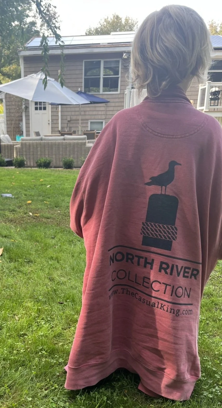 A person with light-colored hair is standing in a backyard, wearing a pink T-shirt with a logo that features a bird sitting on top of a rope-wrapped post, with the text "North River Collection" and a website URL.