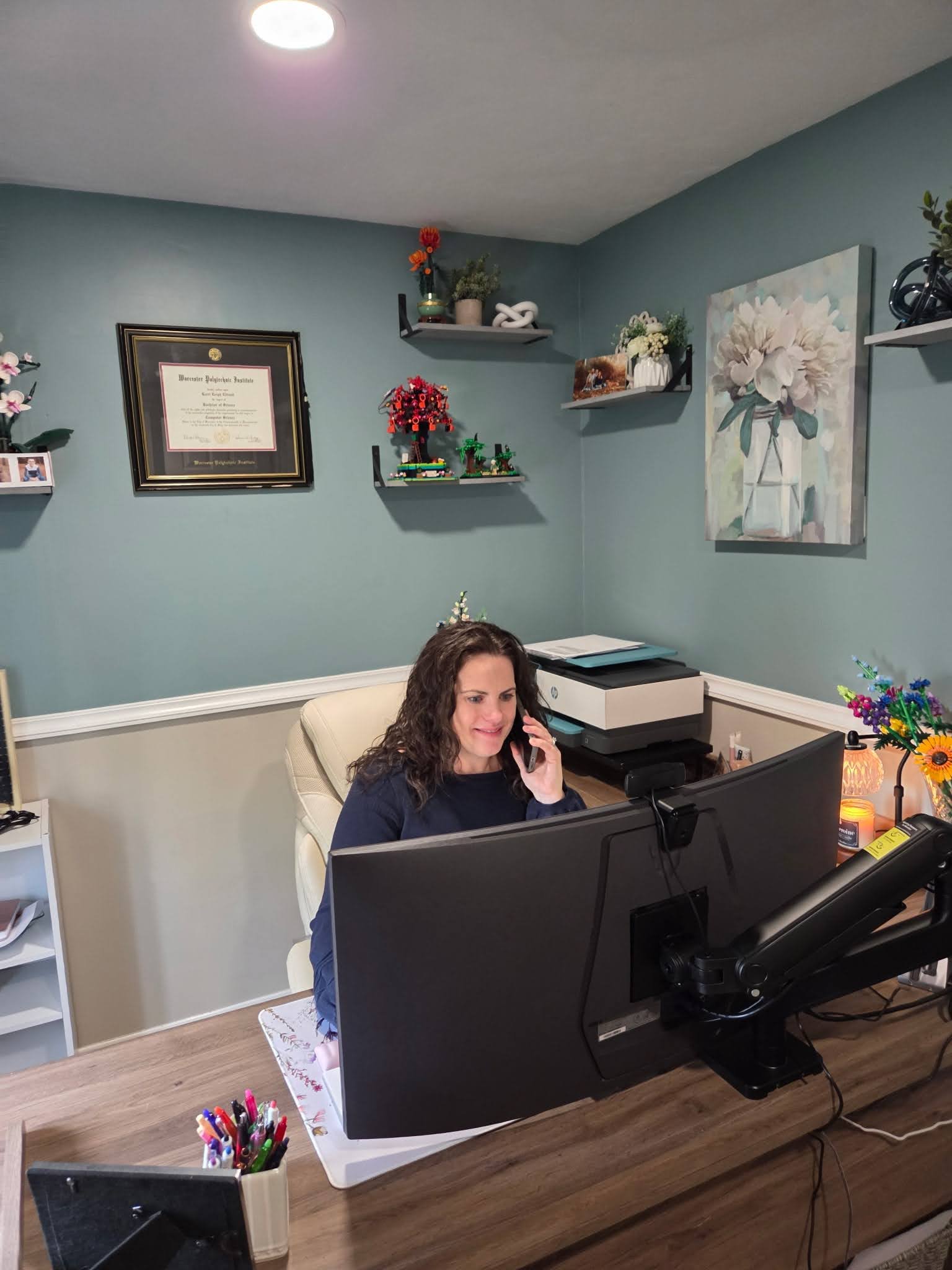 Woman with curly hair sitting at a desk, talking on a phone, working on a computer, in a home office with wall shelves, framed certificate, paintings, and various decorative items.
