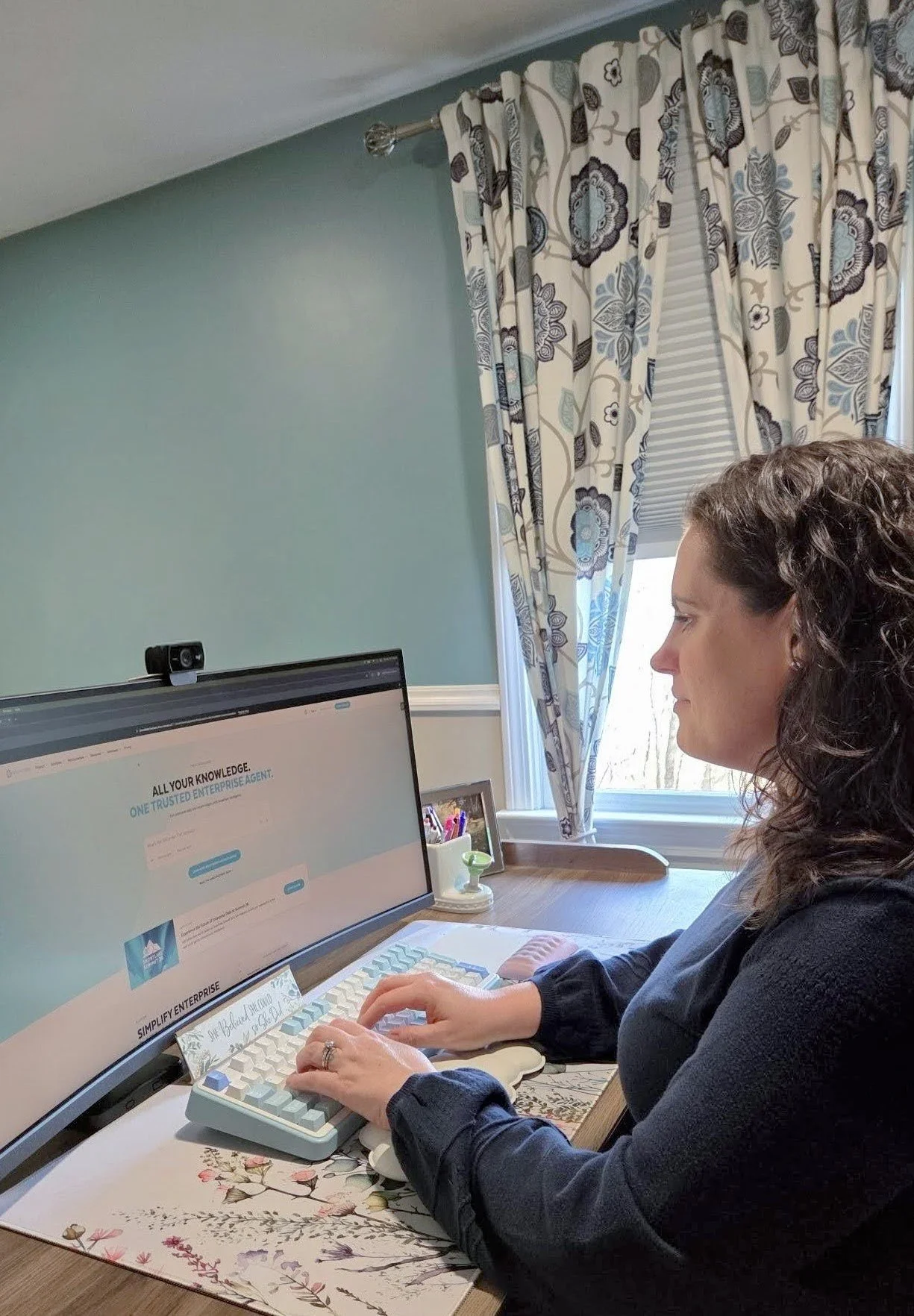 A woman sitting at a desk, working on a computer with a large monitor, in a room with floral curtains and a window, wearing a dark long-sleeved shirt.