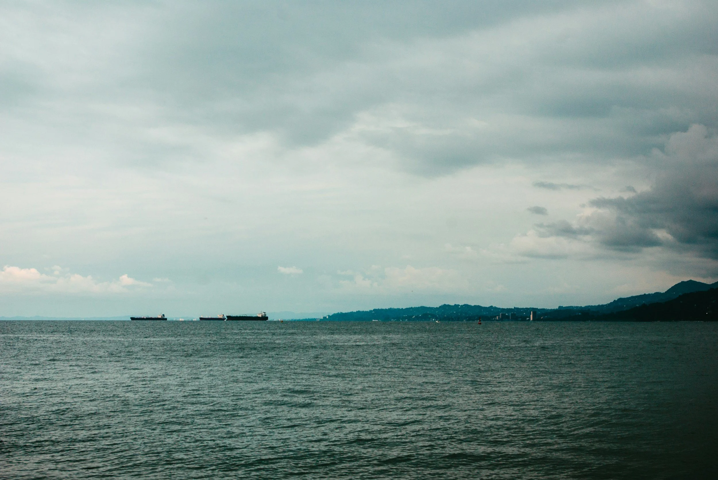 A view of the ocean with three cargo ships in the distance under a cloudy sky, and a distant shoreline with mountains.