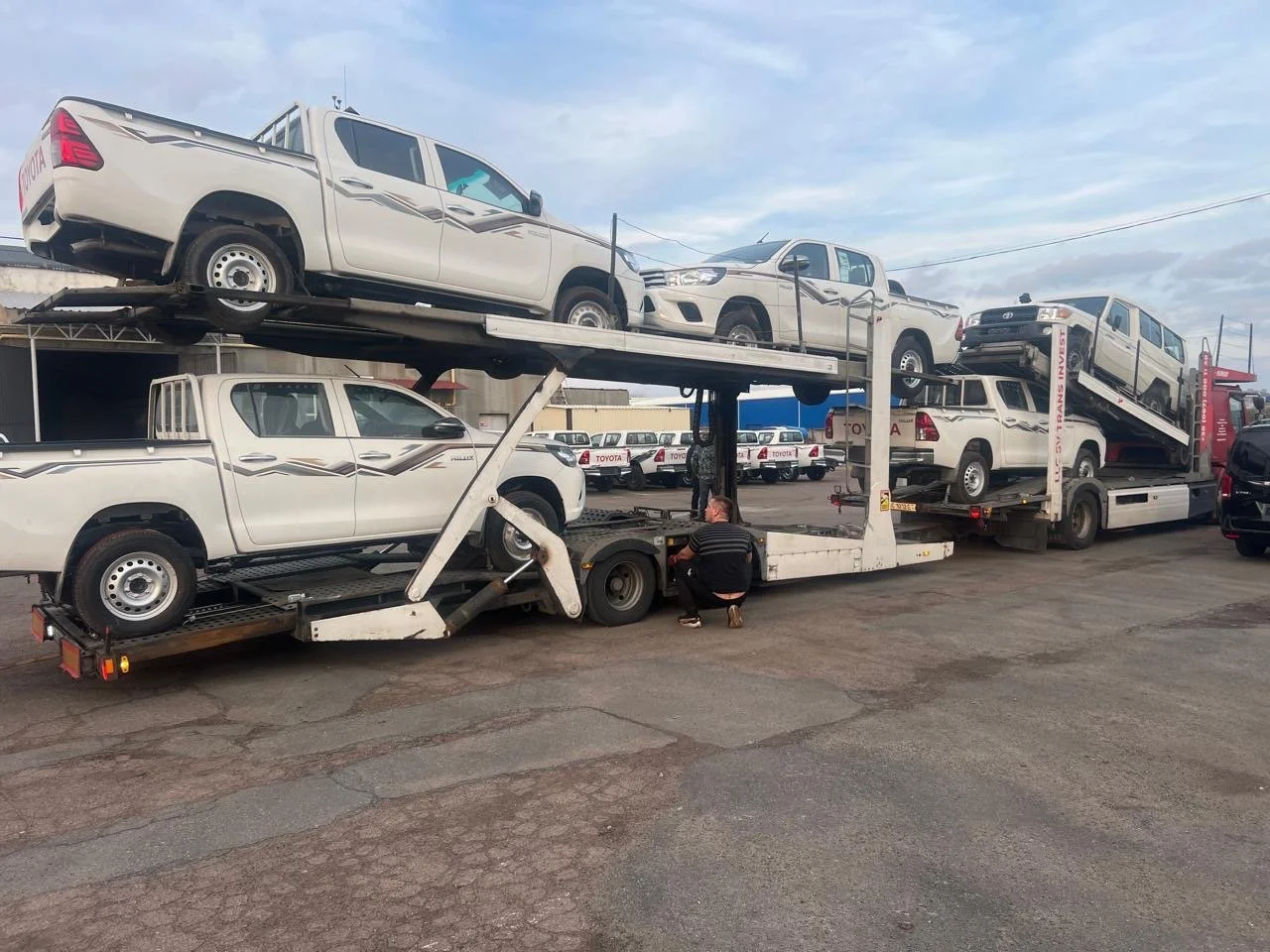A car carrier truck transporting multiple white pickup trucks with black and gray decals parked in an outdoor lot.