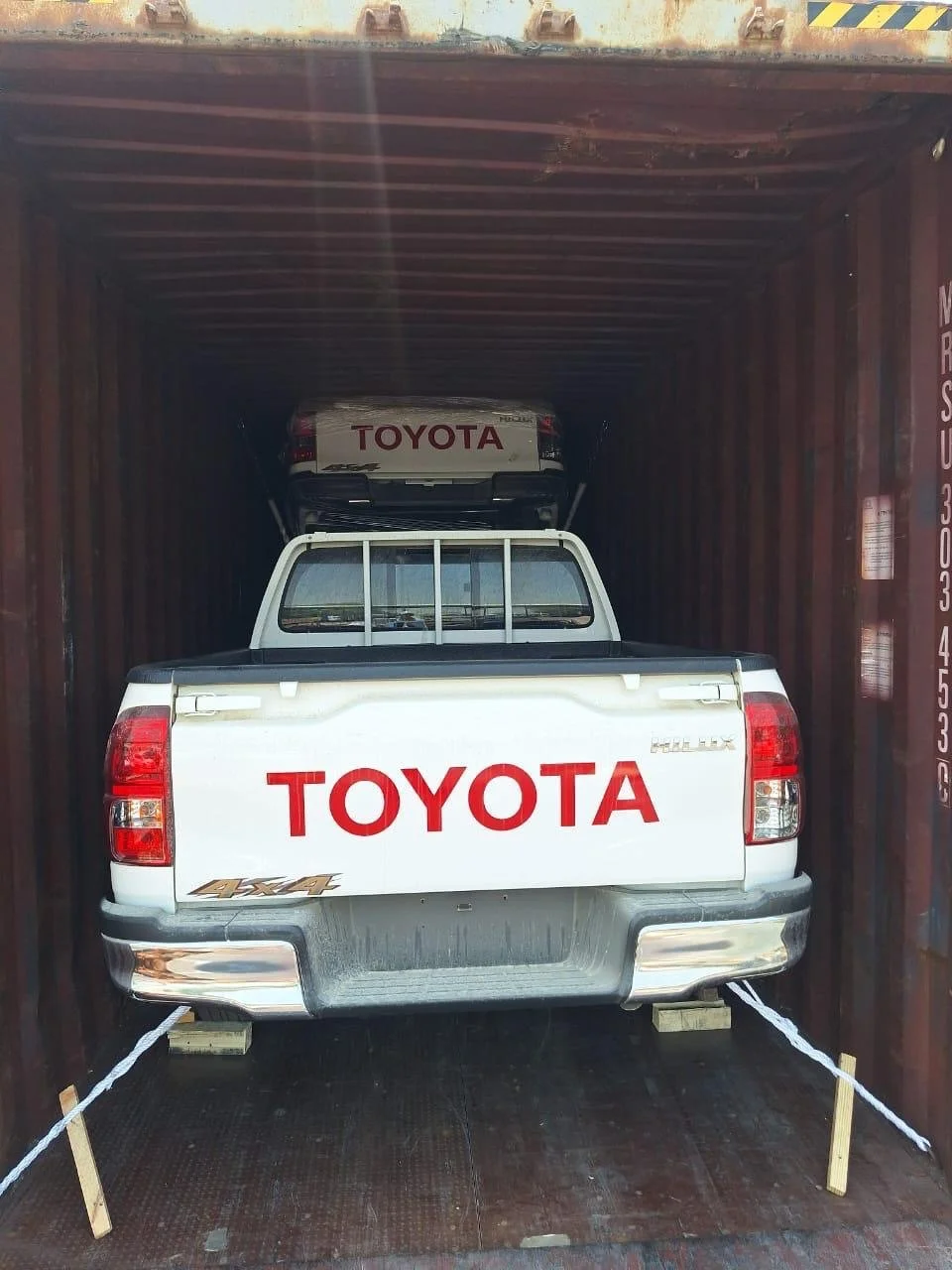 White Toyota pickup truck with large red TOYOTA lettering on the tailgate, secured inside a shipping container with ropes and wooden blocks, with a Toyota car stored on top of the truck.