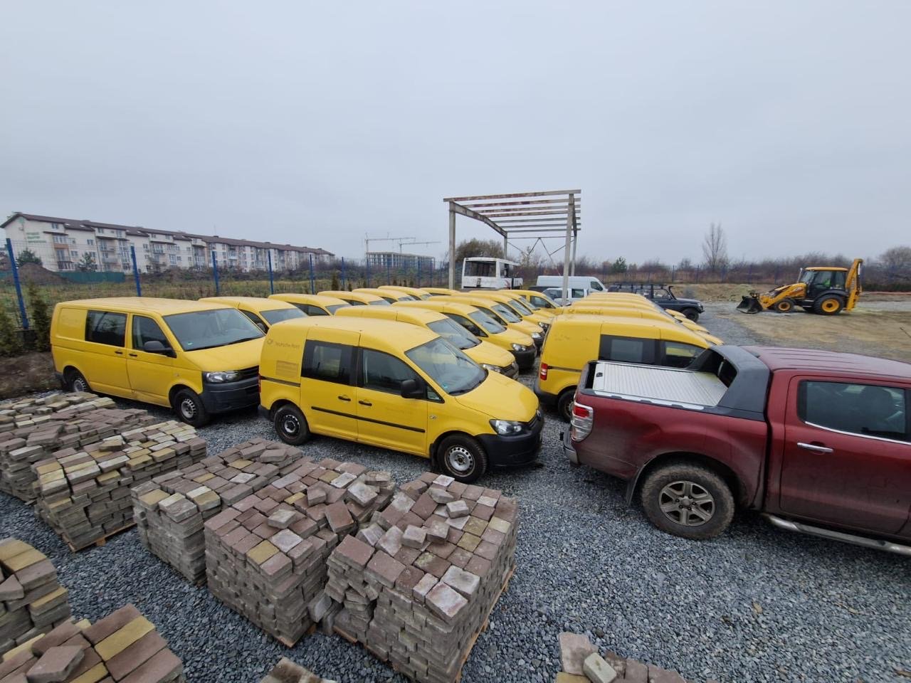 A parking lot with multiple yellow vans and a brown pickup truck, with construction materials and construction equipment in the background.