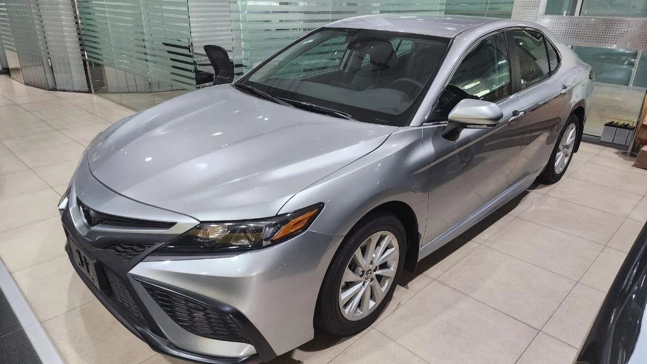 Silver sedan car displayed indoors on a tiled floor, with glass walls and offices in the background.