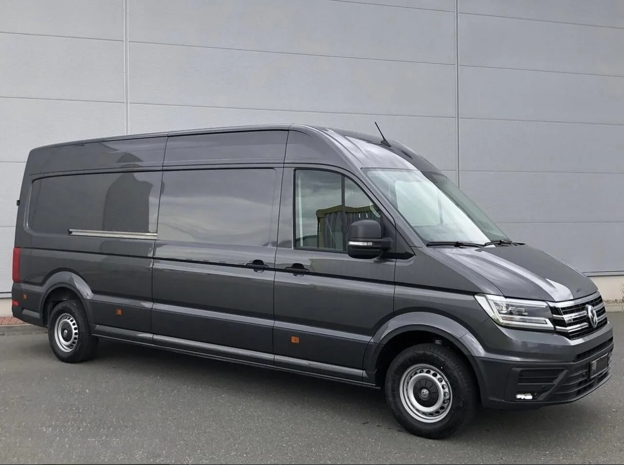 A dark gray Volkswagen commercial cargo van parked in front of a gray industrial building.