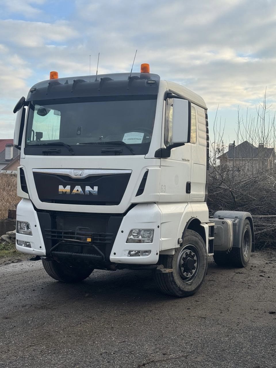 A white MAN semi-truck without a trailer parked outdoors on a dirt ground, with a house and trees in the background under a cloudy sky.