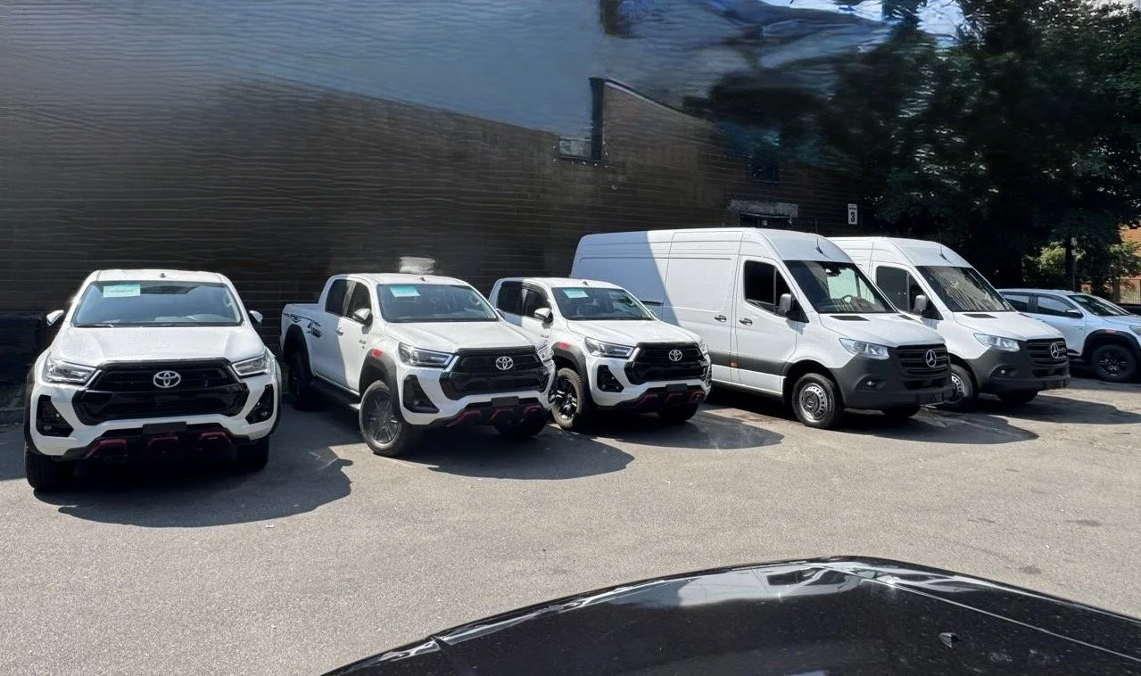 Several white Toyota and Mercedes-Benz commercial vehicles and vans parked in a lot, with a reflective building wall in the background.