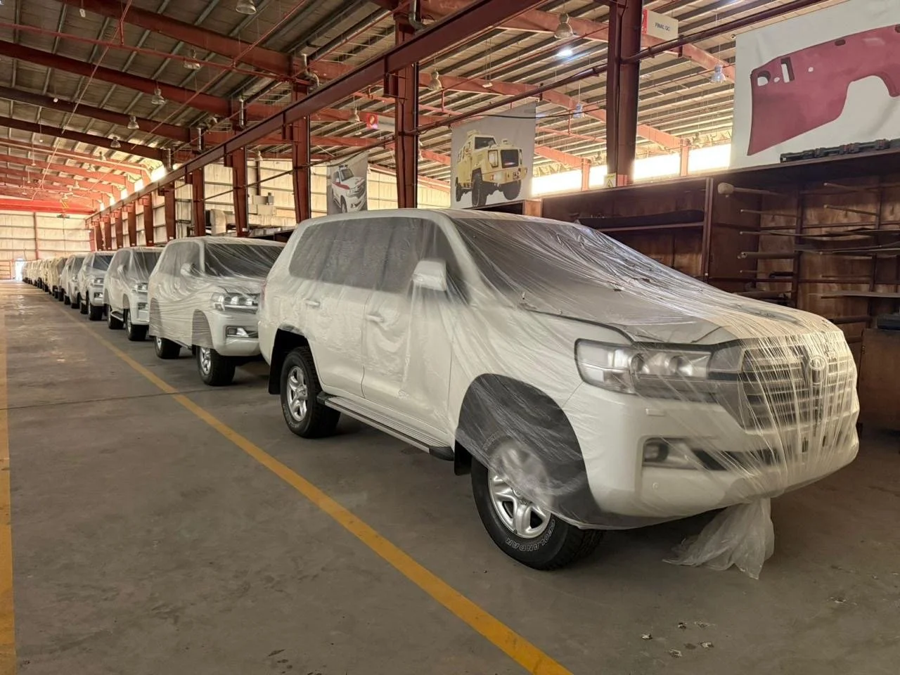 Multiple white cars covered with protective plastic inside a warehouse, with posters of vintage vehicles hanging from the ceiling.