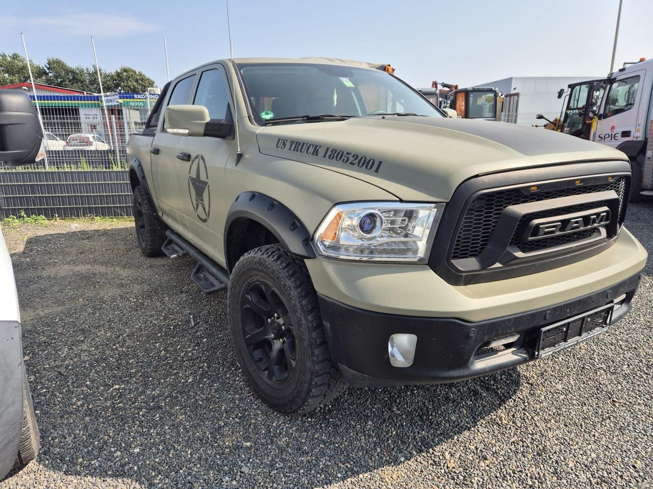 A beige RAM truck with military markings and US military numbers parked on gravel in an outdoor lot.