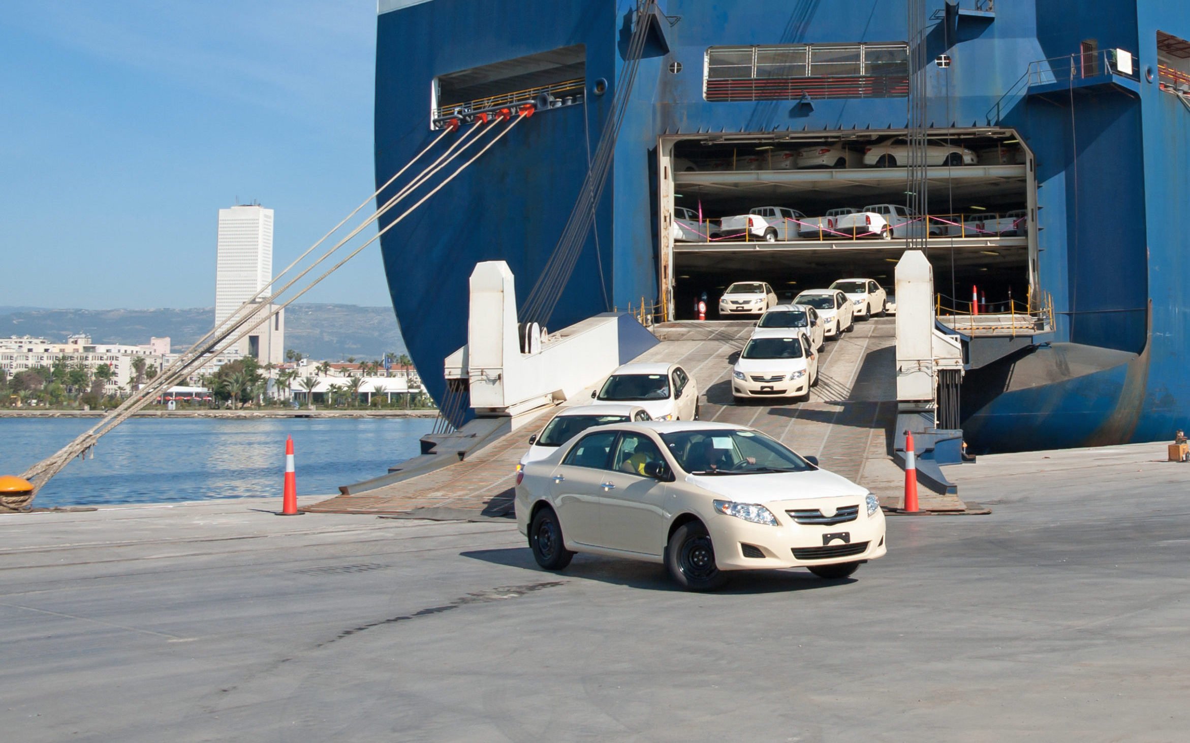 Cars driving off a large ferry boat docked at a harbor, with a city skyline visible in the background.