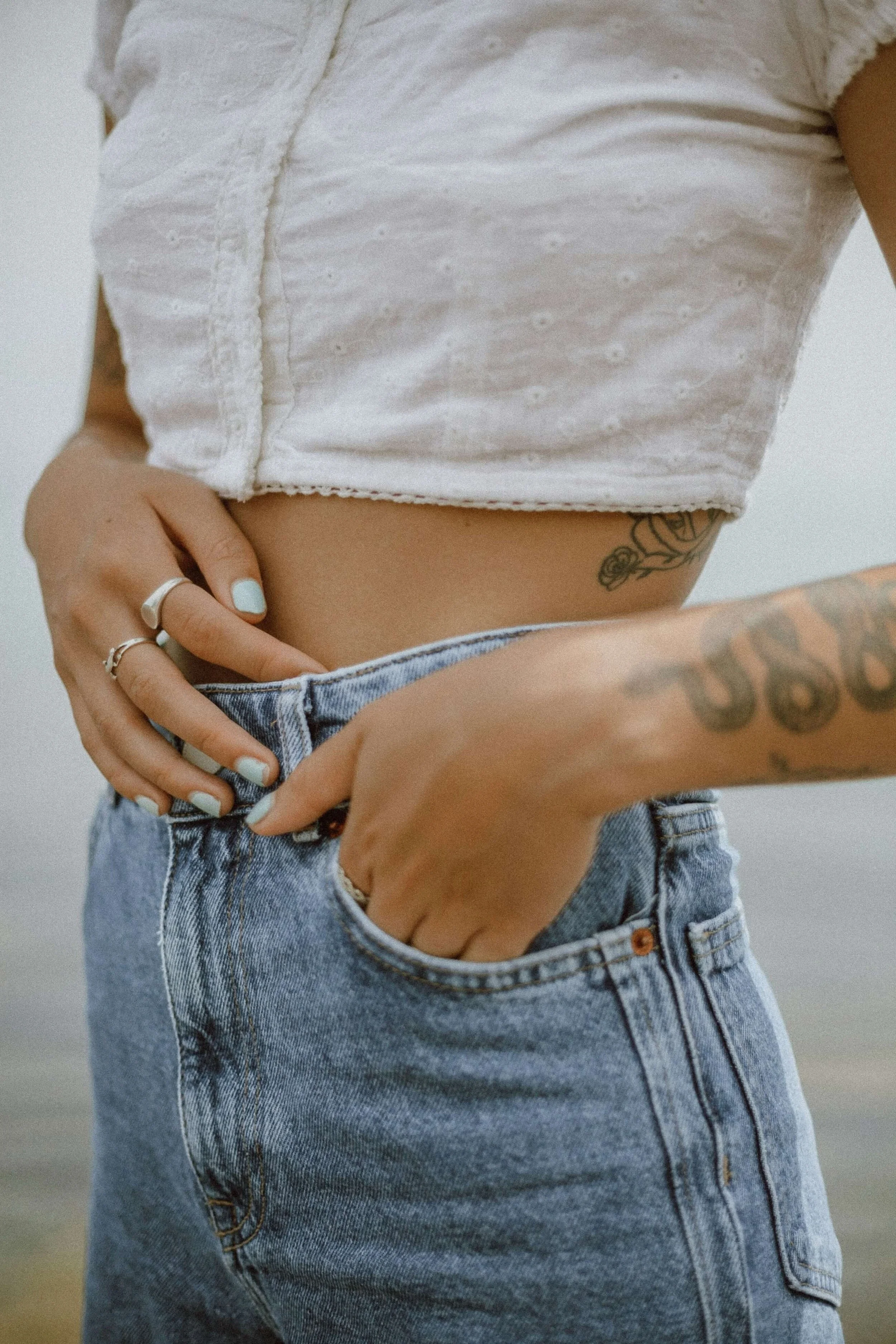 Close-up of a person wearing a white cropped top and high-waisted blue jeans, with tattoos on their hand and side, and rings on their fingers.