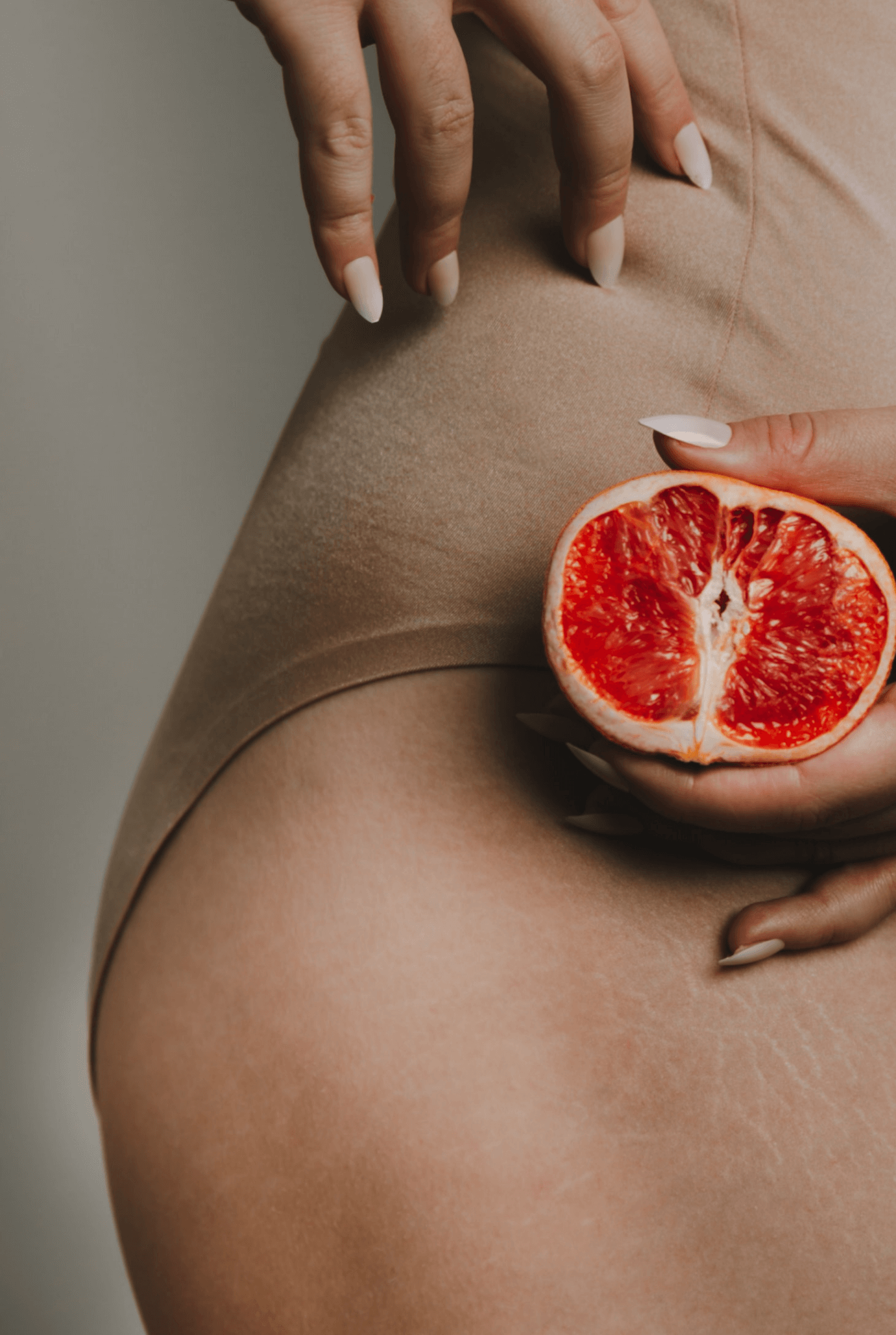 A woman wearing beige shorts holds a halved grapefruit near her thigh.