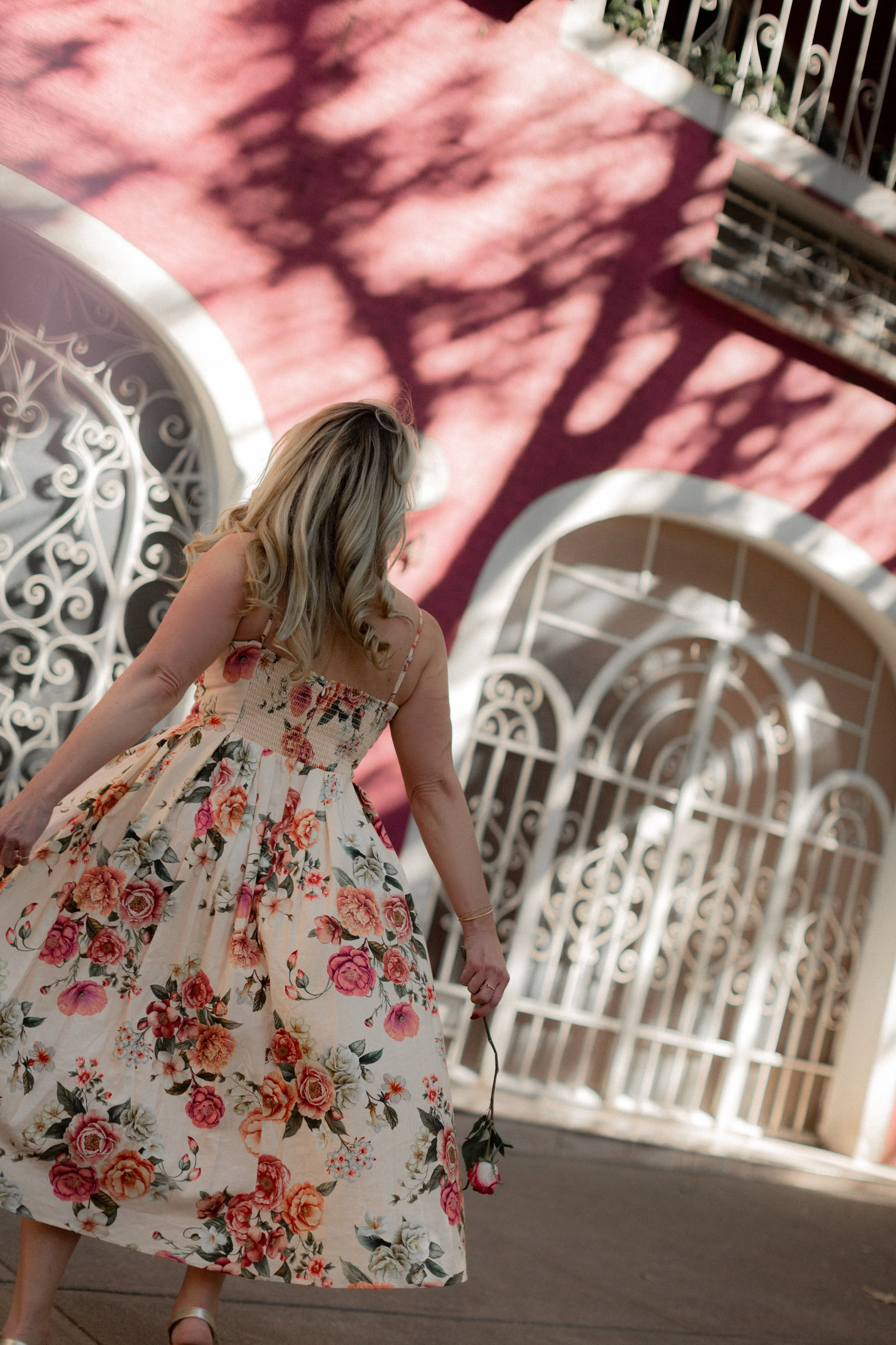 A woman in a floral dress holding a pink rose walking past ornate white gates, with shadows of tree branches on a pink wall.