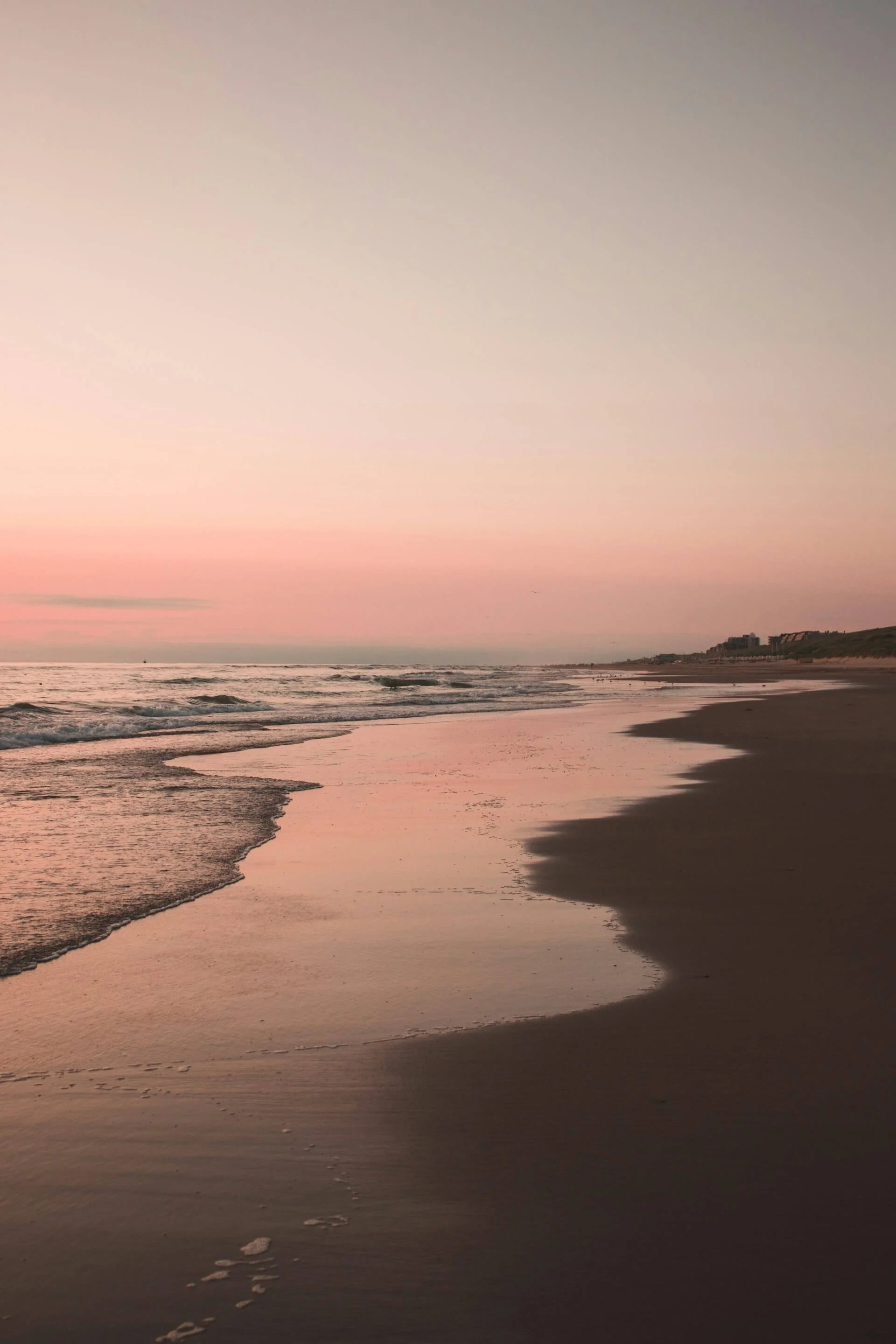 A beach during sunset with calm waves, pinkish sky, and sandy shoreline.