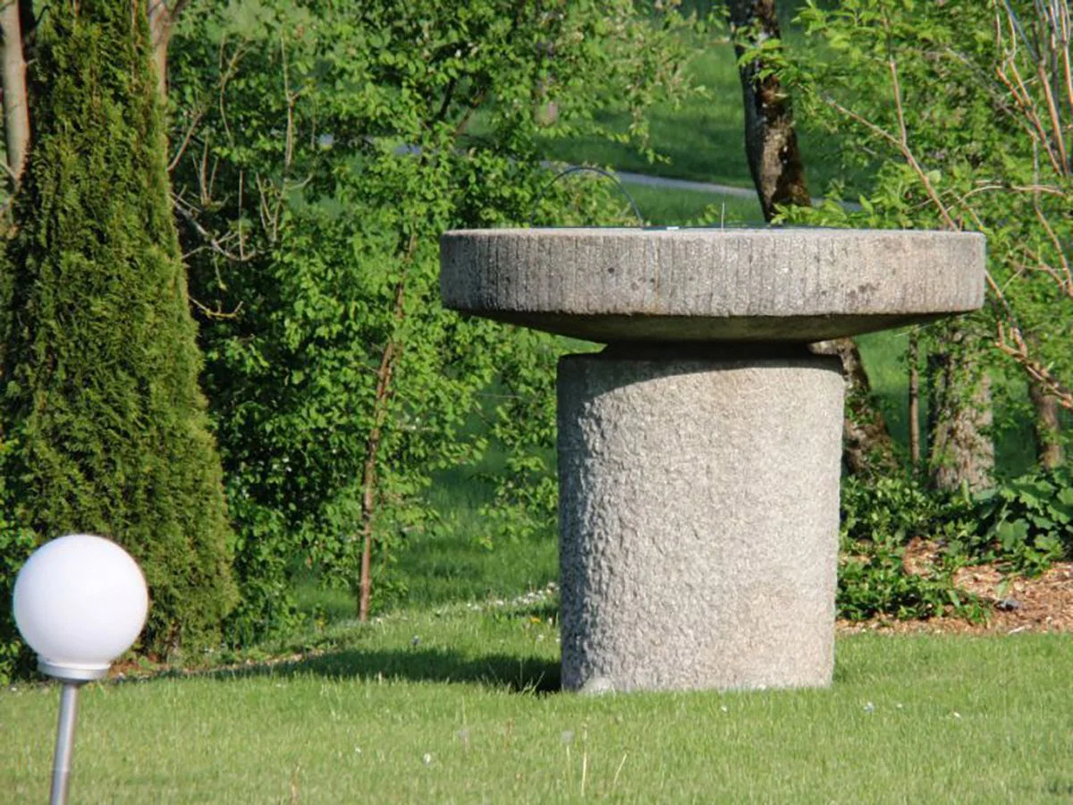 Garten mit einer modernen Skulptur aus Beton, bestehend aus einer runden Platte auf einem zylindrischen Sockel, im Hintergrund Bäume und Büsche.