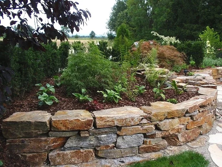 Ein Garten mit einer niedrigen Trockenmauer aus unregelmäßig geformten Steinen, umgeben von verschiedenen Pflanzen und Büschen, mit einem offenen Feld im Hintergrund.
