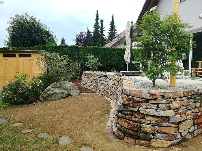 Garten mit Sandkasten, Steinmauer und Pflanzen, Haus im Hintergrund, bewölkter Himmel.