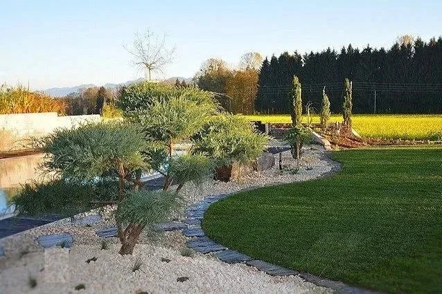 Garten mit grünem Rasen, Kieswegen und einigen Sträuchern, Bäumen und Hecken, mit dem Himmel im Hintergrund.