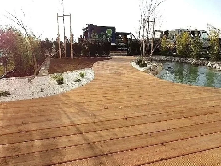 Ein Holzsteg führt zu einem kleinen Teich mit Steinen am Ufer, im Hintergrund sind Bäume — gartengestaltung
