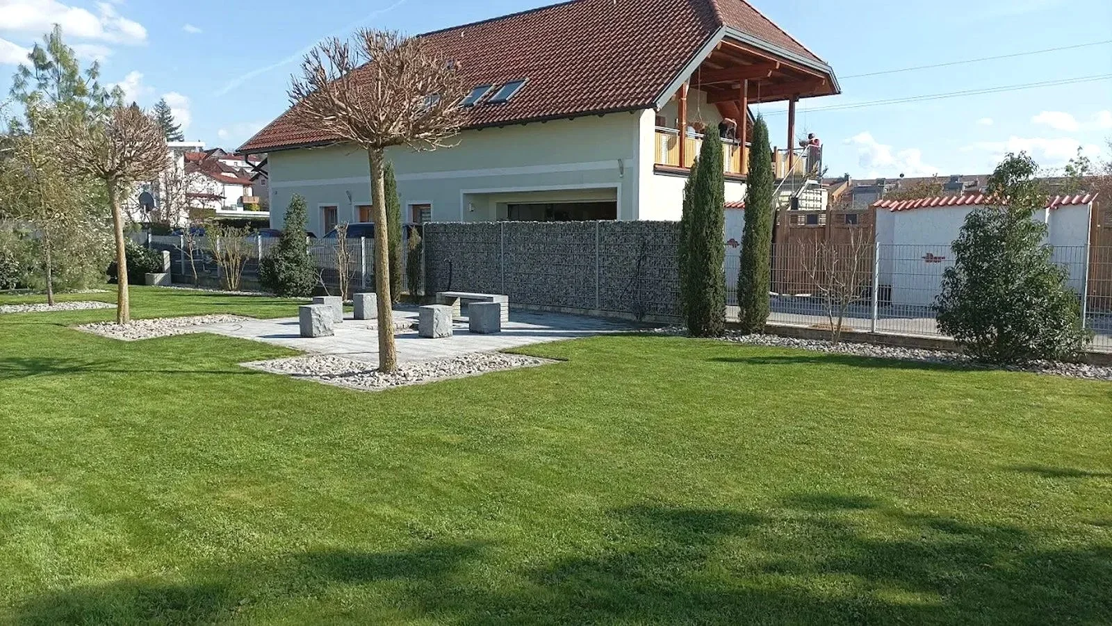 Garten mit grünem Rasen, vier Bäume, gepflasterte Sitzbank mit Steinen, Haus mit Balkon im Hintergrund, bewölkter Himmel.