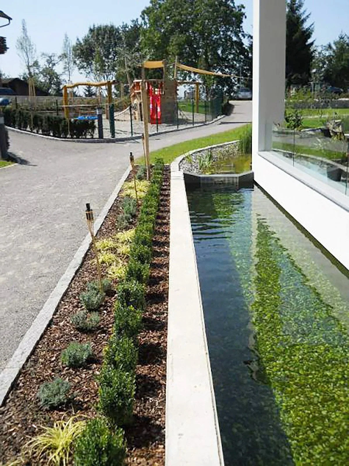 Ein Garten mit einem kleinen Wasserlauf neben einem Gebäude. Im Hintergrund ein Spielplatz mit Klettergerüst und Rutsche, umgeben von Bäumen.