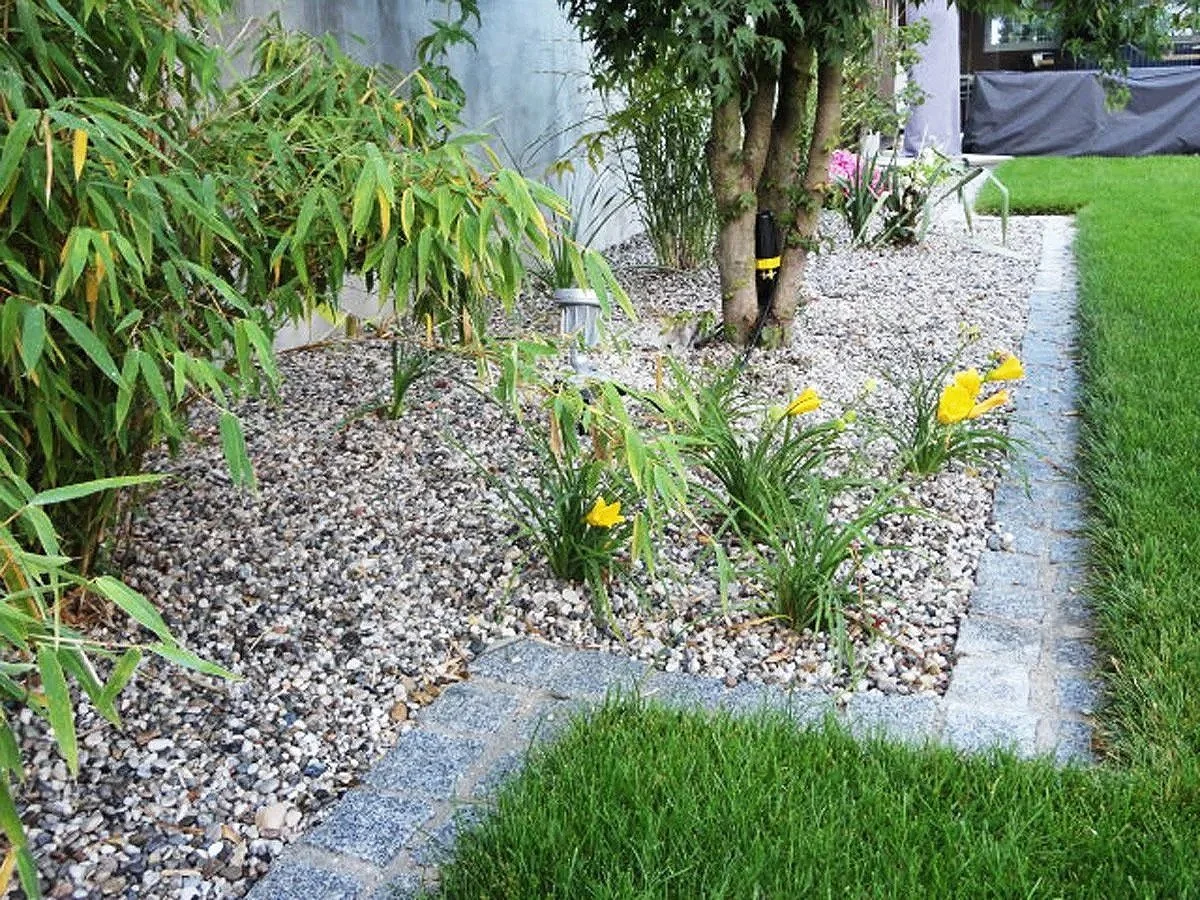 Garten mit Kieselsteinbedeckung, grünem Rasen, kleinen Büschen, bunten Blumen und einem Baum.