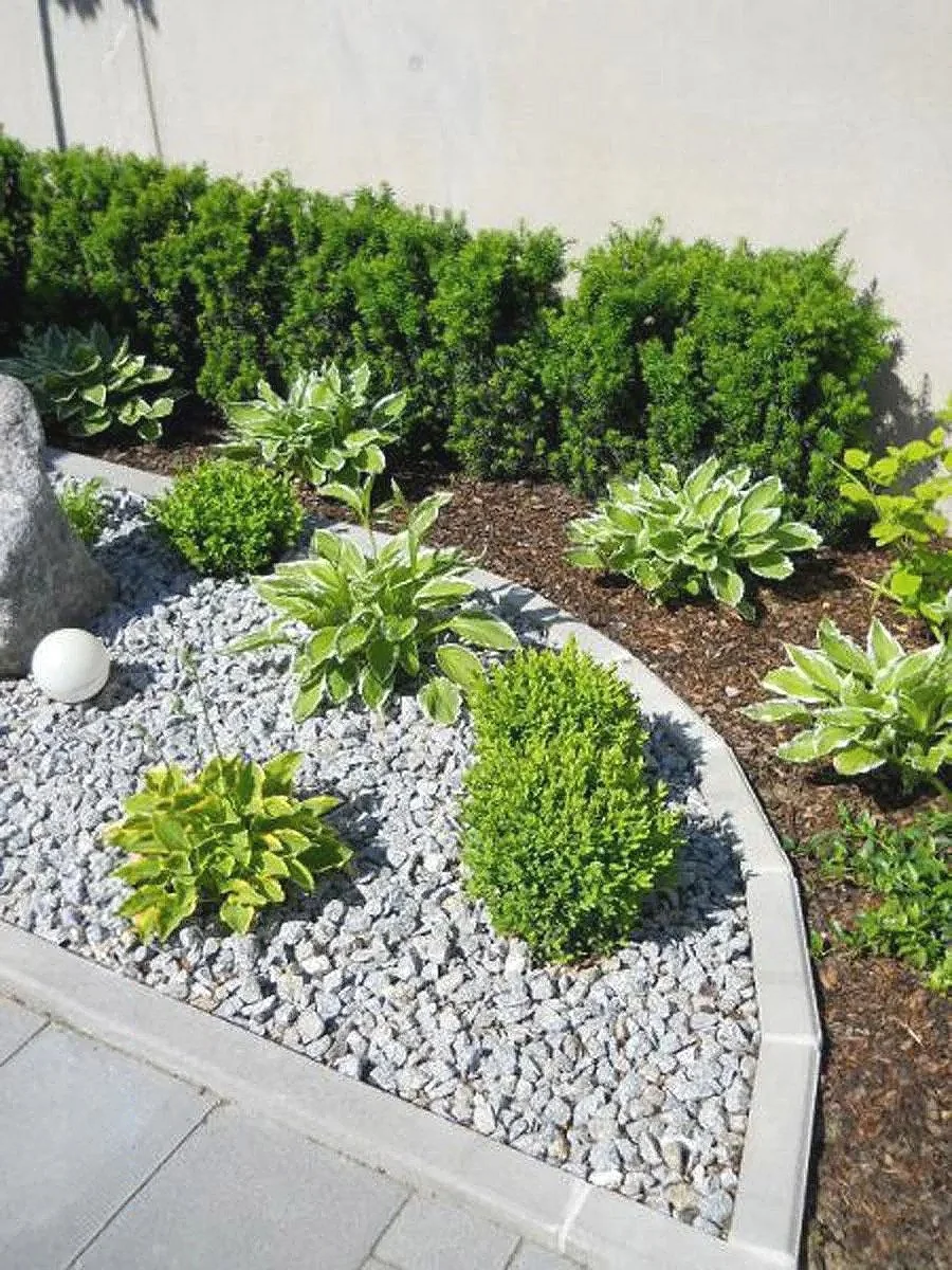 Garten mit grünen Sträuchern, weißen Steinen und einer weißen Gartenlampe, umgeben von einer Steinmauer und einem weißen Zementboden.
