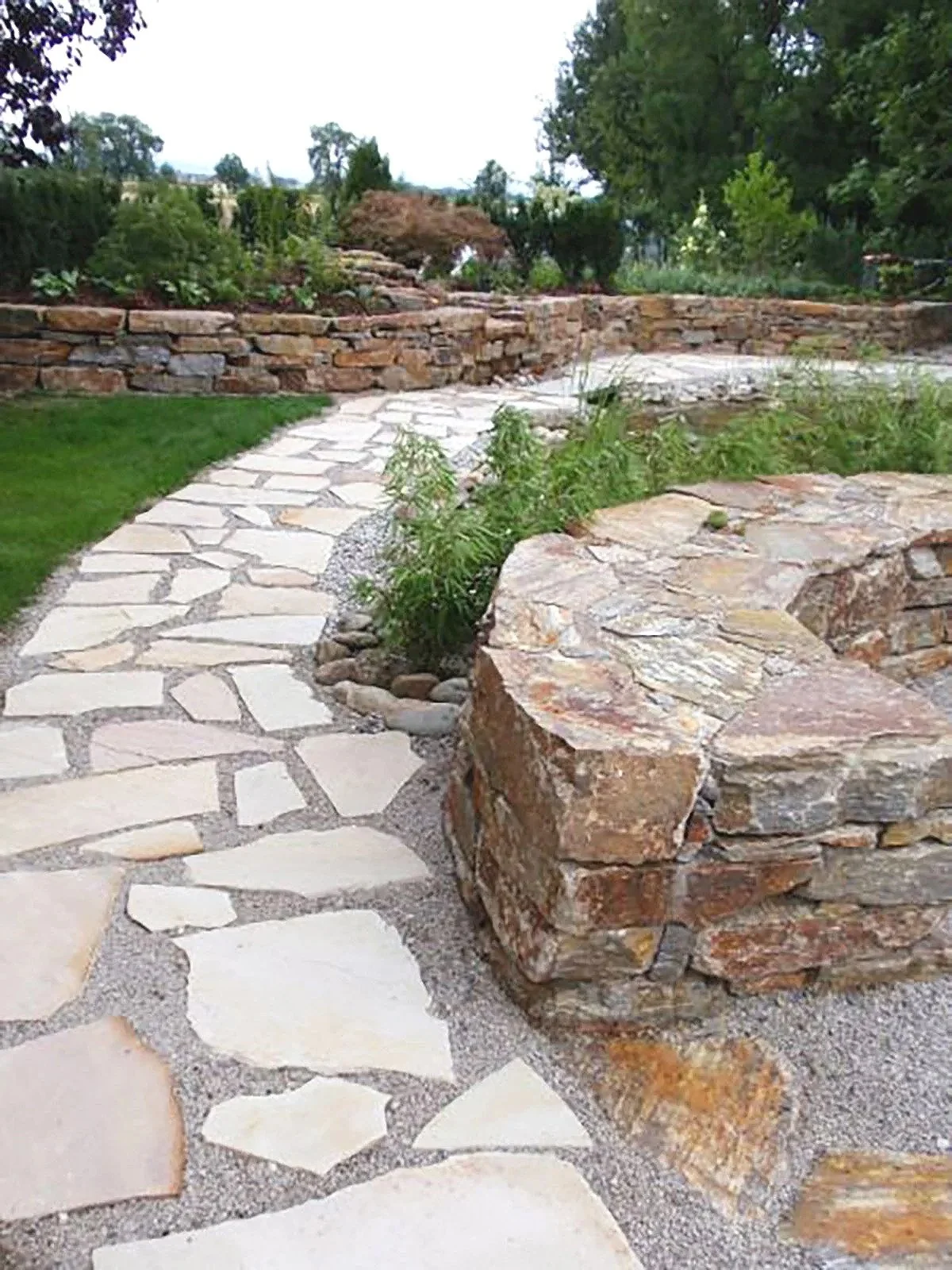 Ein Gartenweg aus unregelmäßig geschnittenen Steinen, umrahmt von einem Terrassendickem mit großen Steinen und kleinen Pflanzen, befindet sich in einem gepflegten Garten mit Bäumen und Sträuchern.