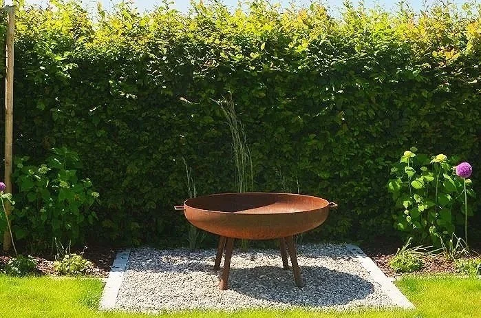 Eine rustikale, runde Feuerschale auf drei Beinen in einem Garten mit grünen Hecken und bunten Blumen.