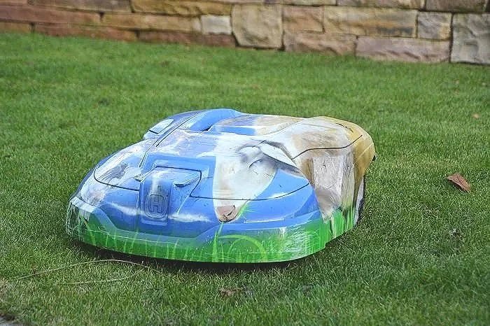 Ein gehäkelter Rasenmähroboter, der wie eine Hund, eine Katze und einen Fuchs aussieht, auf einem grünen Rasen mit Steinmauer im Hintergrund.