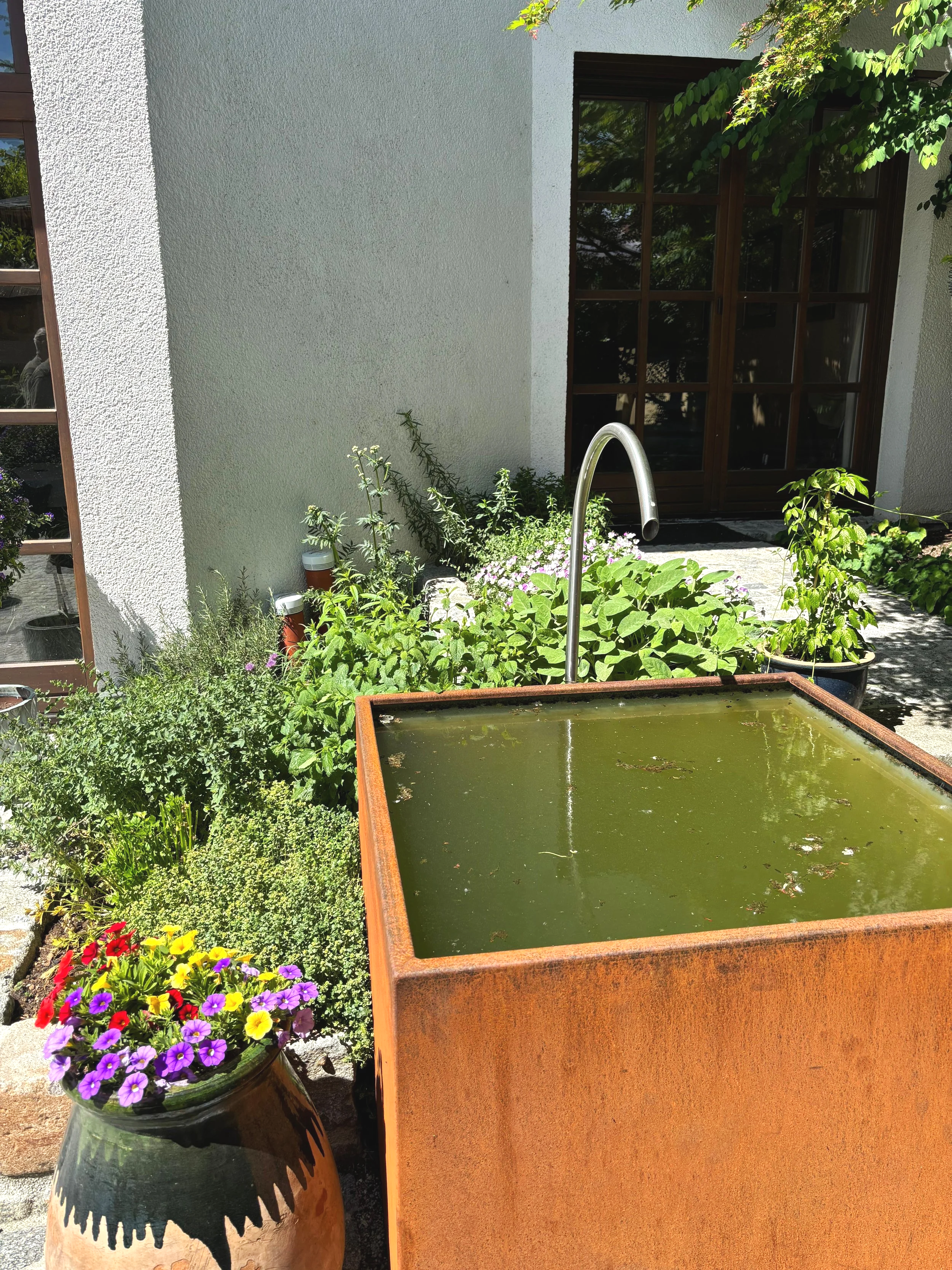 Ein Garten mit einem hohen, rustikalen Brunnen mit Wasser und einem Wasserhahn in der Mitte, umgeben von Blumen und grünem Pflanzen vor einer weißen Hauswand mit Holztür.