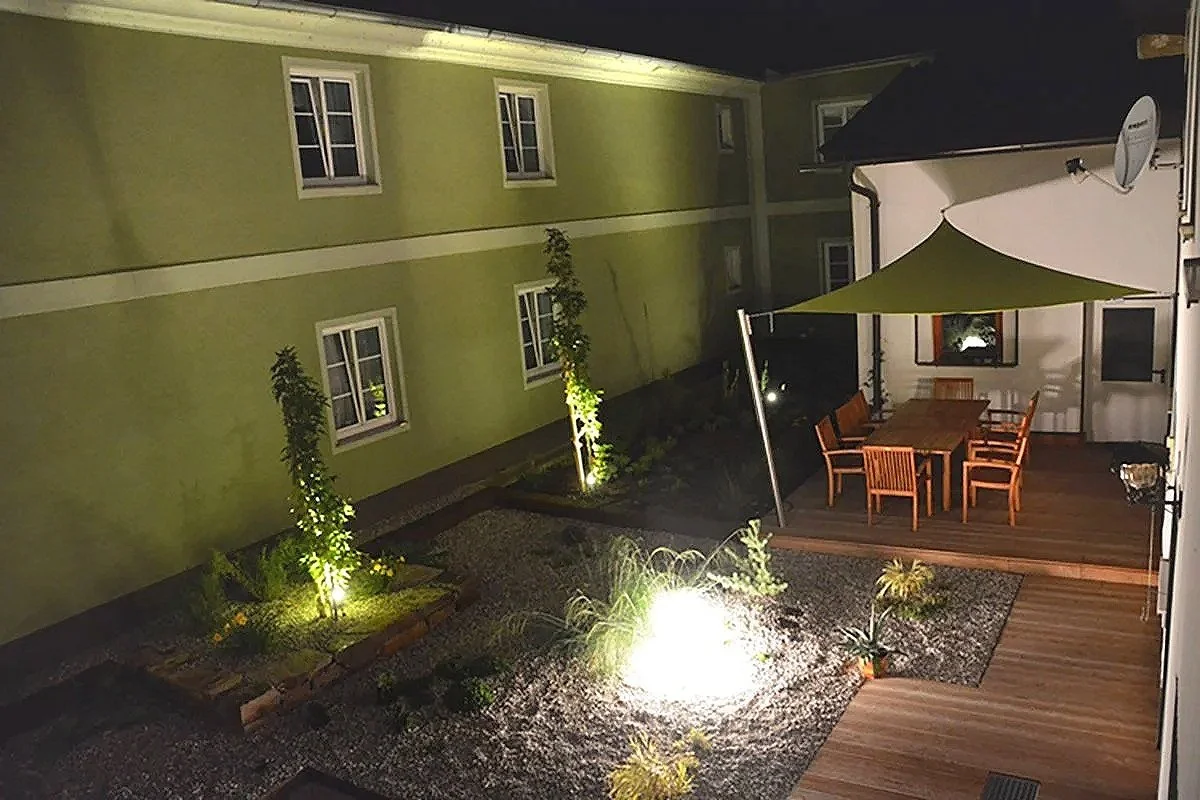 Ein Garten mit einer Holzterrasse, Gartenmöbel, Sonnenschirm, bepflanzten Blumenbeeten und Hausfenstern bei Nacht, beleuchtet von Außenleuchten.