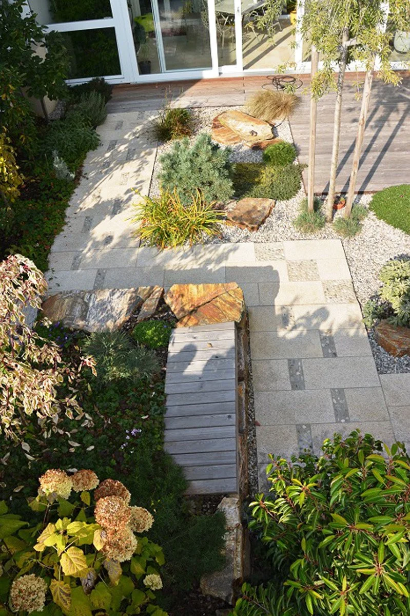 Garten mit Steinen, Pflanzen und Holzweg vor einem modernen Haus mit großen Glasfenstern.