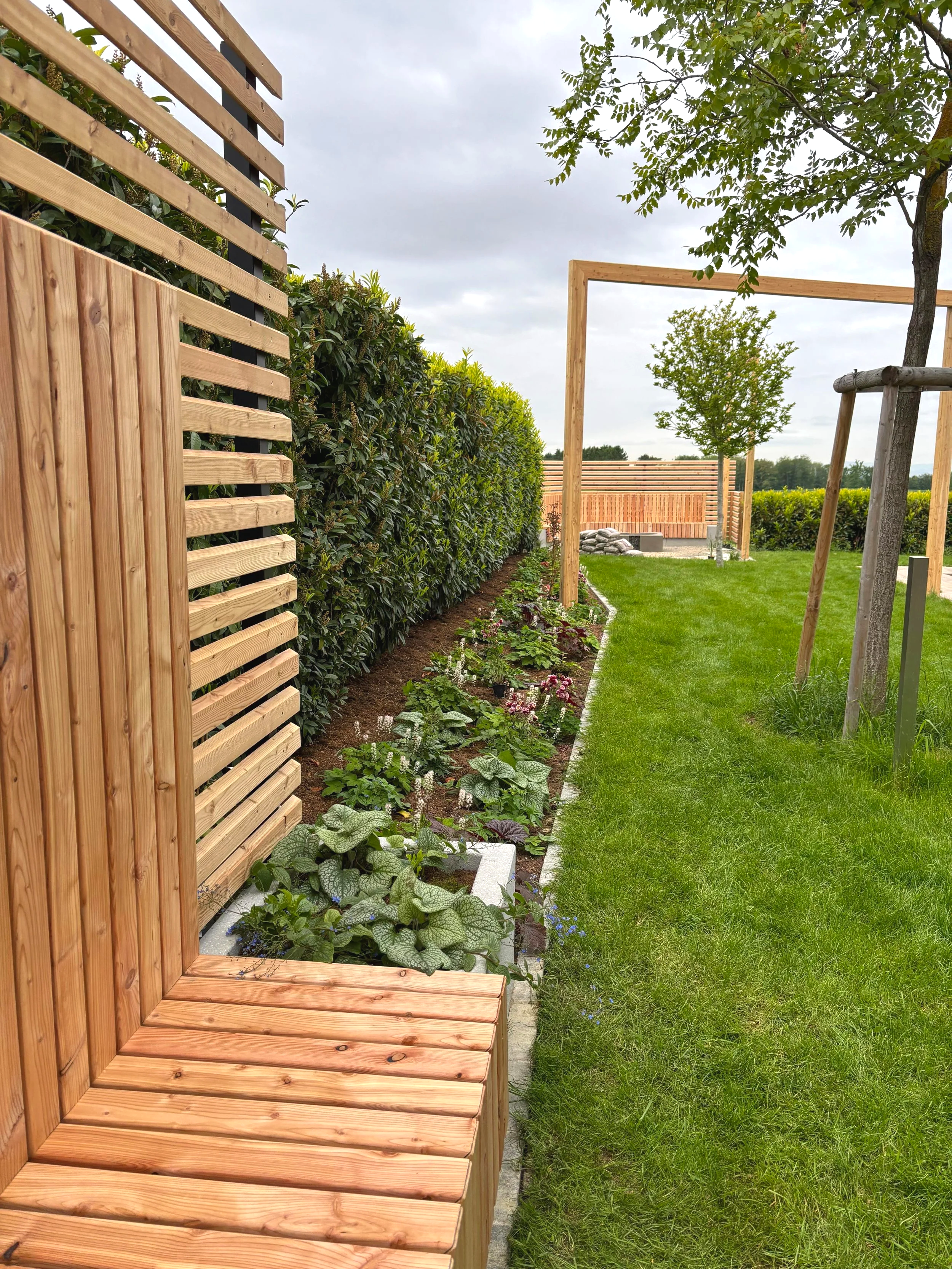 Garten mit Holzgestaltungen, Blumenbeet, grünem Rasen und Bäumen unter bewölktem Himmel.