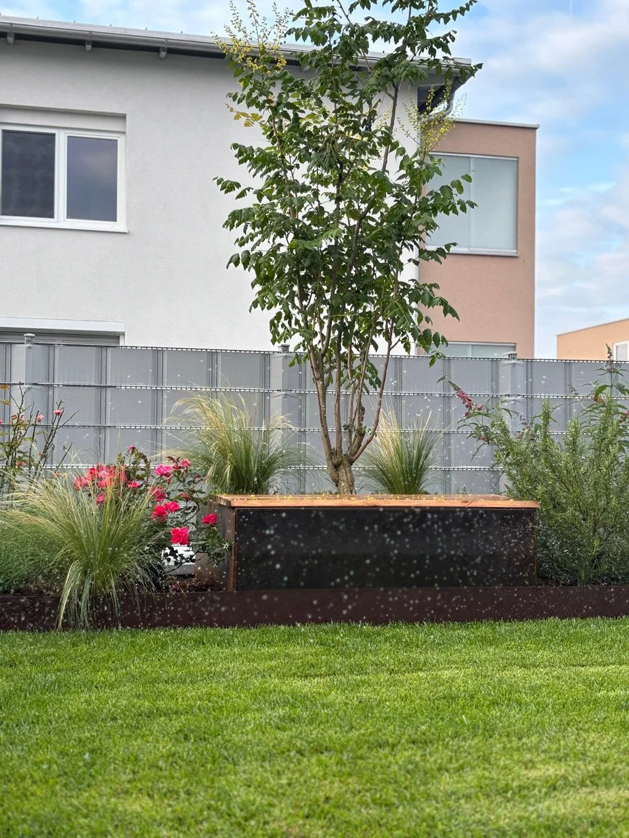 Garten mit Blumen, Bäumen und Rasen vor einem Mehrfamilienhaus