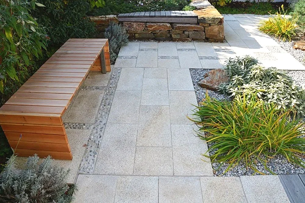 Ein Garten mit Steinterrasse, einer Holzbank, Rasen, verschiedenen Pflanzen und einem kleinen Steinbrunnen.