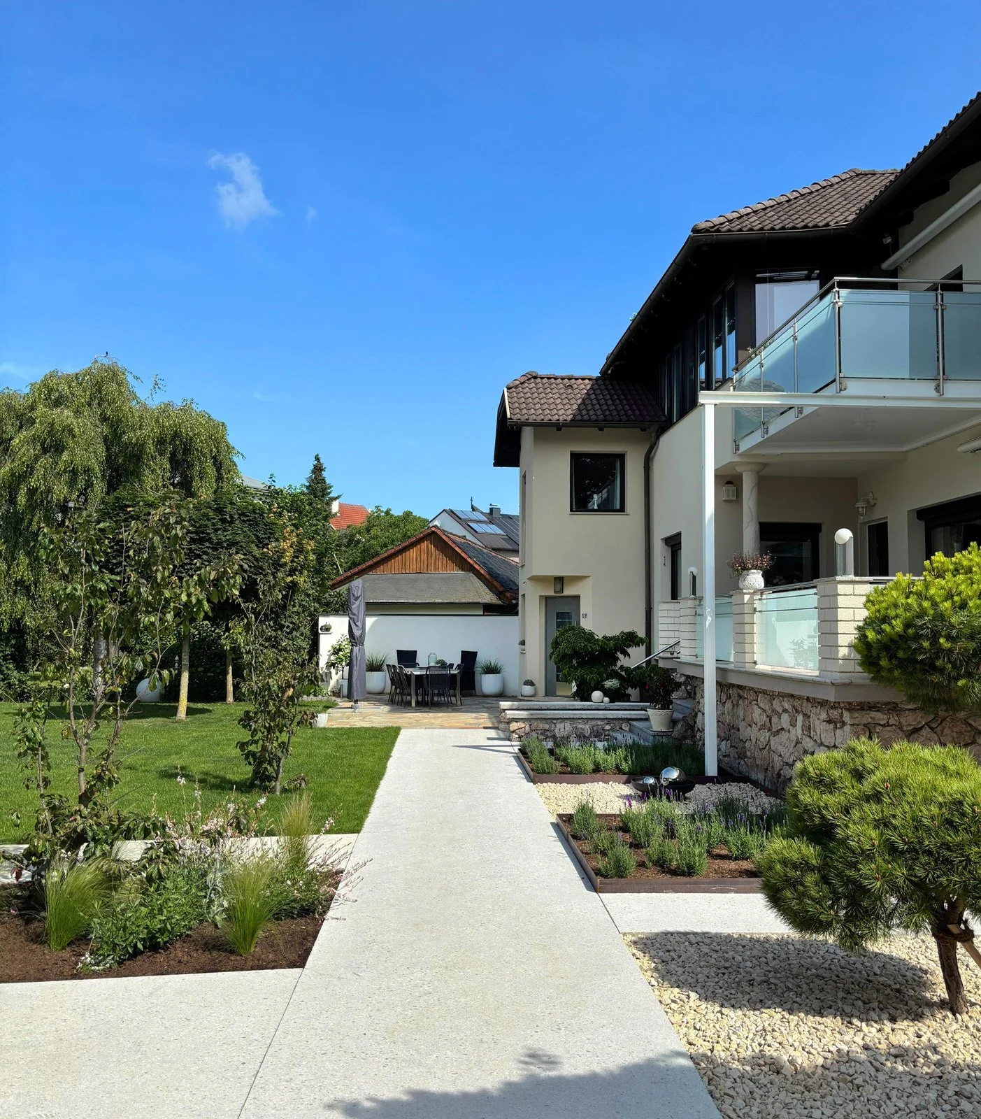 Garten mit Gehweg, grünem Rasen, Bäumen und Sträuchern vor modernem Hauses mit Terrasse und Balkon bei sonnigem Himmel. Gartengestaltung Niederösterreich