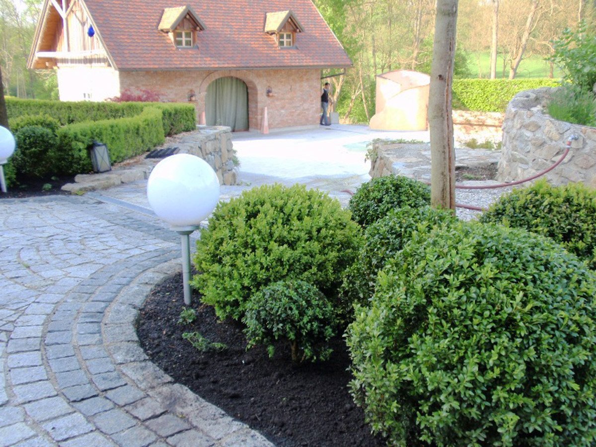 Garten mit gepflastertem Weg, runden Sträuchern, weißen Kugelleuchten und einem Haus im Hintergrund