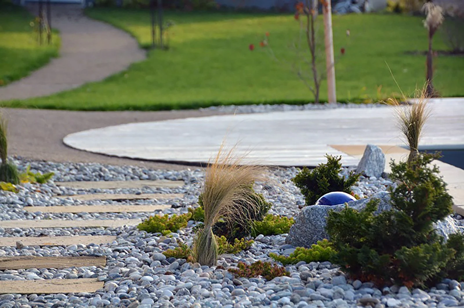 Garten mit Steinen, Pflanzen und einer gepflasterten Weg, im Hintergrund ist eine Wiese und eine schmale Teerstraße.