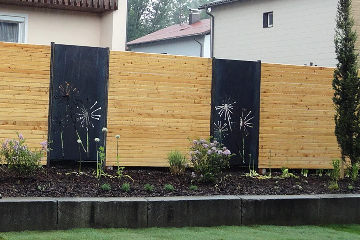 Gartenzaun aus Holz mit schwarzen Toren mit dandelion-Designs, umgeben von blühenden Pflanzen und ein Haus im Hintergrund.