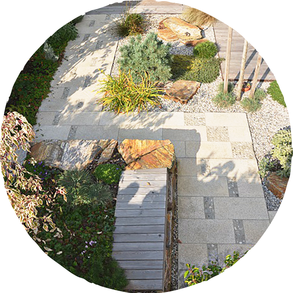 Ein Gartenweg mit Pflastersteinen, umgeben von Steinen und Pflanzen, inklusive kleiner Bäume und Stauden.