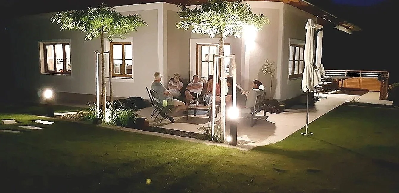 Menschen sitzen auf einer Terrasse eines Hauses bei Nacht, umgeben von Bäumen und Gartenbeleuchtung.