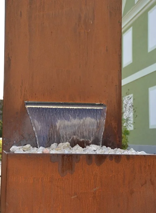 Eine moderne Brunnenanlage aus rostigem Metall mit Wasser, das über eine kleine Wasserfallkante fließt, umgeben von weißen Kieselsteinen.