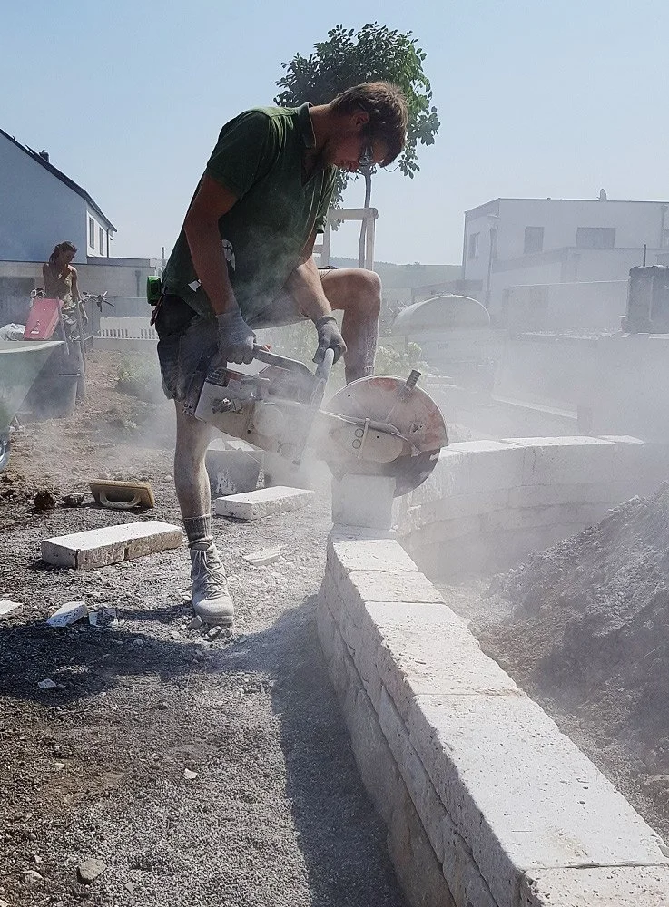 Ein Mann beim Mauern mit einem elektrischen Zementmischer, bei heißem Sonnenlicht, in einem Gartenbereich.