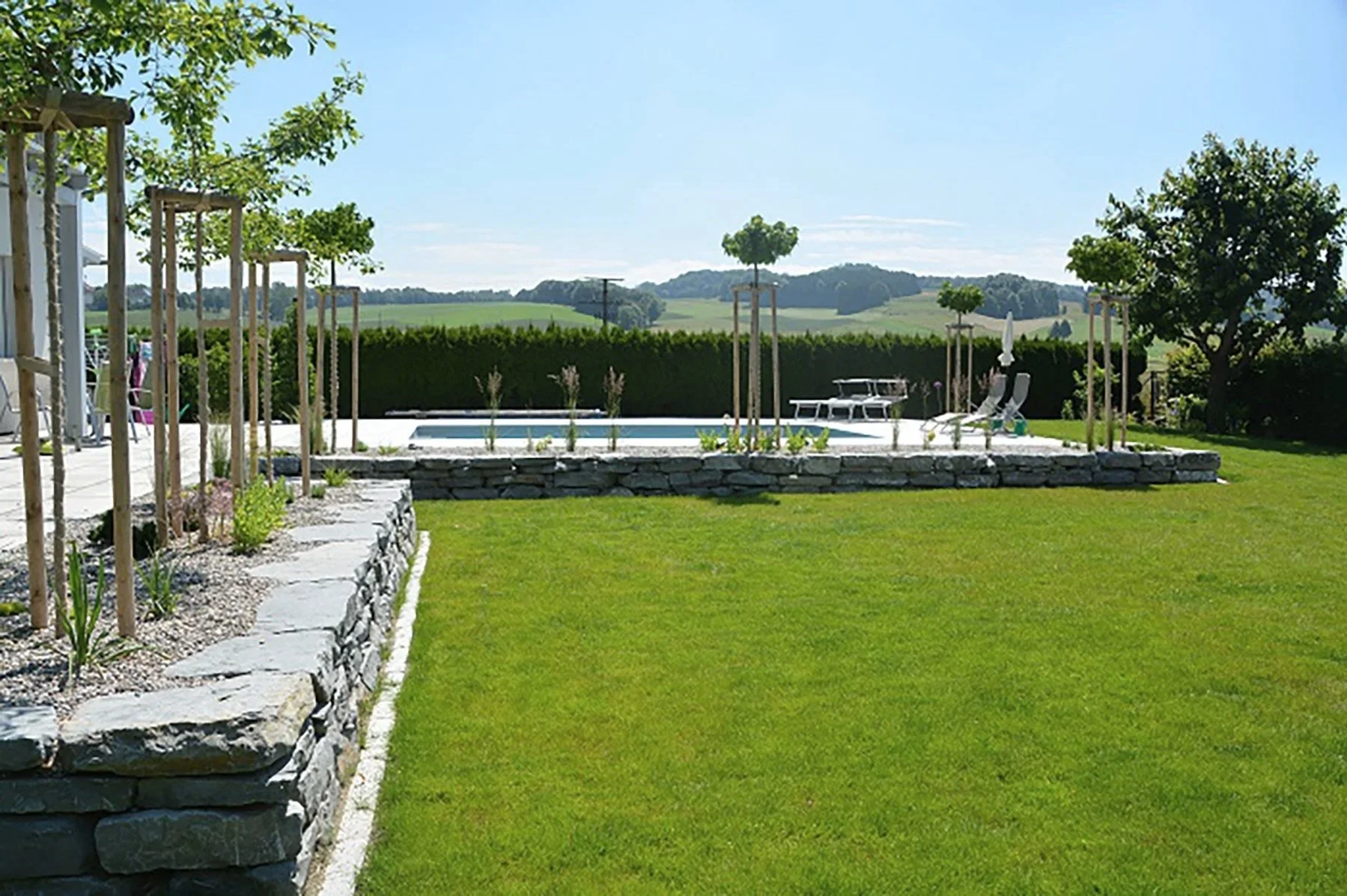 Gartengestaltung, die Gärtnerin — Garten mit grünem Rasen, kleinen Bäumen mit Stützpfählen, einer Steinterrasse, einem Pool, Liegestühlen und einem Boot davor, mit Hügeln im Hintergrund bei sonnigem Himmel.