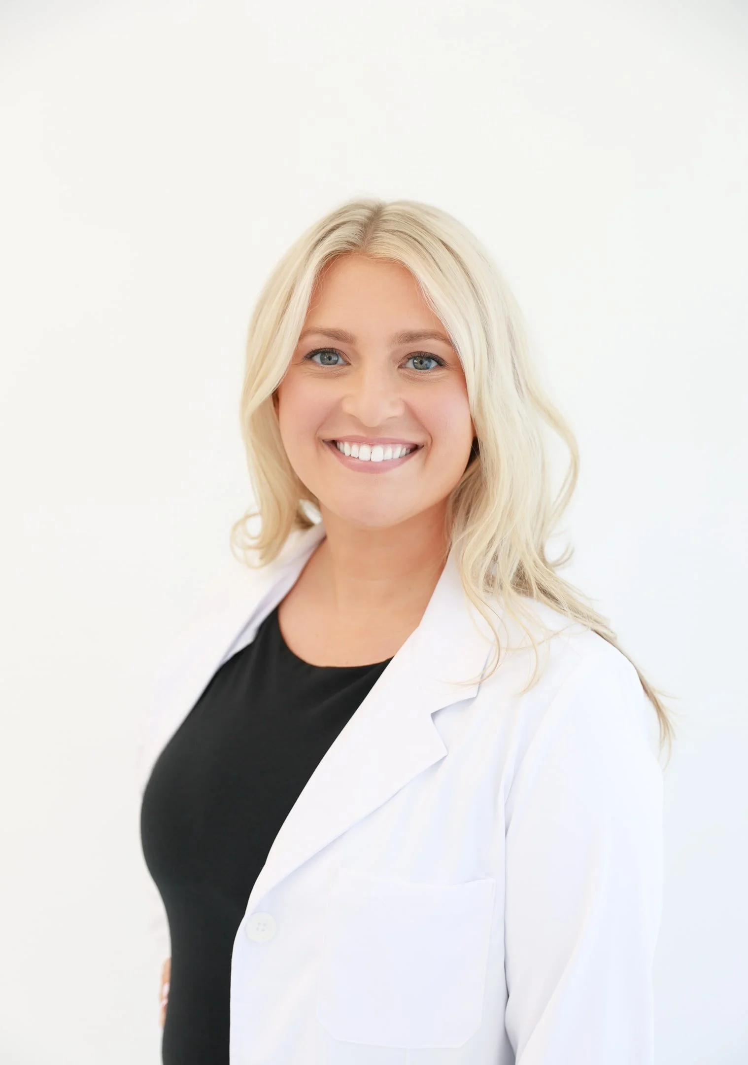 A smiling blonde woman with blue eyes, wearing a black top and a white lab coat, standing against a plain white background.